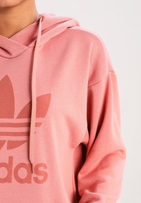 Růžová mikina s kapsou na klokany a předním potiskem se třemi pruhy a trojlístkovým logem. Směs bavlny, měkká textura.