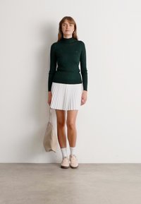 Femme se tenant contre un mur blanc, portant un col roulé vert foncé, une jupe plissée blanche, des chaussettes blanches, des baskets beiges et tenant un sac à main beige.