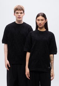 Μαύρα πλεκτά υφασμάτινα μπλουζάκια με κοντά μανίκια, με υφαντό σχέδιο και ριμπ λαιμόκοψη· φορεμένα με αντίστοιχο μαύρο, φαρδύ παντελόνι.