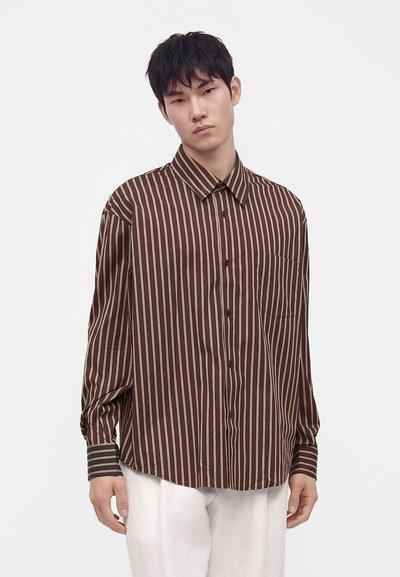 Jeune homme portant une chemise marron ample à rayures beiges verticales et un pantalon blanc, debout devant un fond clair uni.