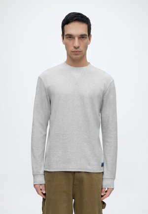 Homme aux cheveux courts et foncés portant une chemise gris clair à manches longues et un pantalon vert olive sur un fond blanc uni.