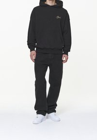 Svart huvtröja med en texturerad design, framficka och guld broderad logotyp; matchad med svarta jeans och vita sneakers.