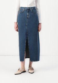 Denim midi-skjørt med frontsplitt, høy midje, fem lommer og subtile stikninger. Brukt med lysebeige flate sko.