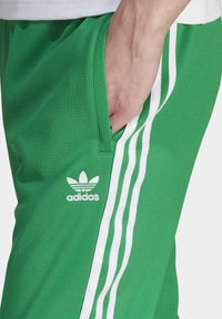 Gröna atletiska byxor i mjukt tyg med vita sidsstreck och fickor. Har Adidas-logotypen i vitt framtill på vänster sida.