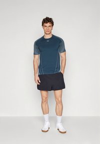 Lühikeste varrukatega sportlik särk tumedassinises toonis helesiniste aktsentidega, kombineeritud mustade lühikeste pükste ja valgete tossudega, esitatud neutraalsel taustal.