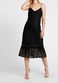 Svart spets midi klänning med tunna axelremmar, V-ringning och ett volangkjol. Texturen är intrikat med ett blommönster, kompletterad med silverfärgade klackar.