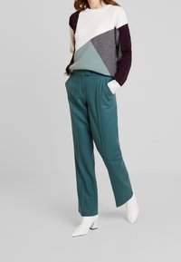 Vrouw in teal hoge taille rechte broek, witte laarzen en een kleurbloktrui met witte, grijze, teal en donkerpaarse panelen.