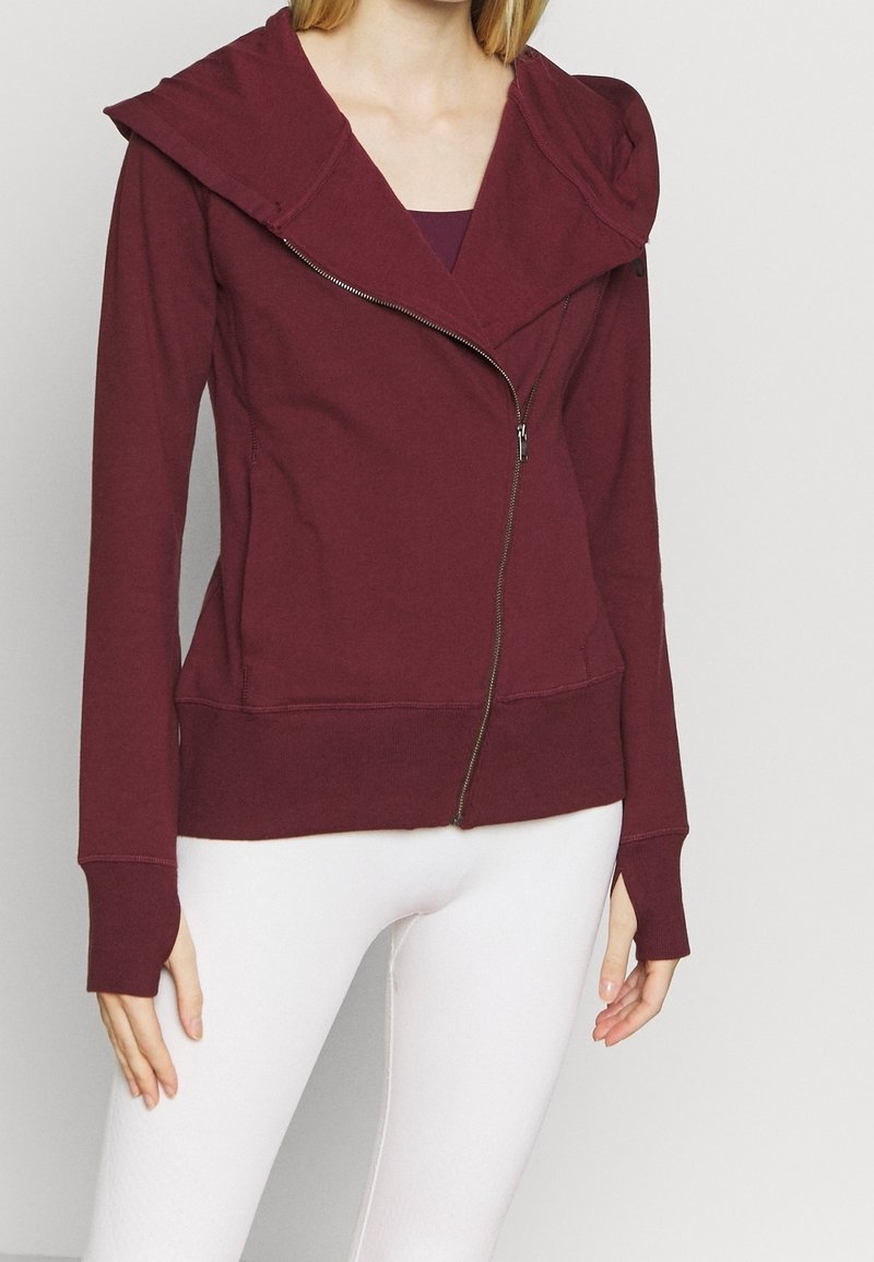 Bordeaux zip hoodie med en stor hætte, tætsiddende manchetter og en glat tekstur. Har en skråtgående lynlås og en afslappet silhuet.