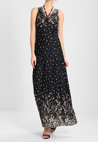 Czarna maxi sukienka w kwiaty z marszczonym dekoltem, zdobiona małymi białymi kwiatami i brązowym wzorem kwiatowym u dołu. Gładka tekstura materiału.