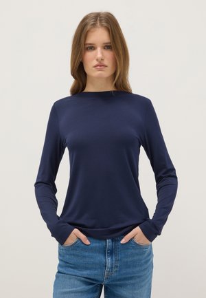 Junge Frau mit langen blonden Haaren, trägt ein dunkelblaues Langarmshirt und blaue Jeans, Hände in den Taschen, neutraler Hintergrund.