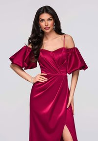 Robe en satin fuchsia avec épaules dénudées, manches bouffantes ; comprend un corsage ajusté, des détails drapés et une fente haute sur une cuisse.