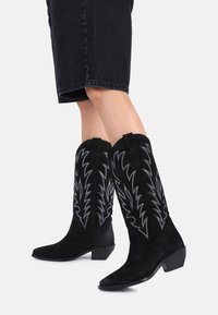 Sorte semskede cowboystøvler med broderte bladmønstre, spisse tær og lave blokkerte hæler, parret med denimshorts.