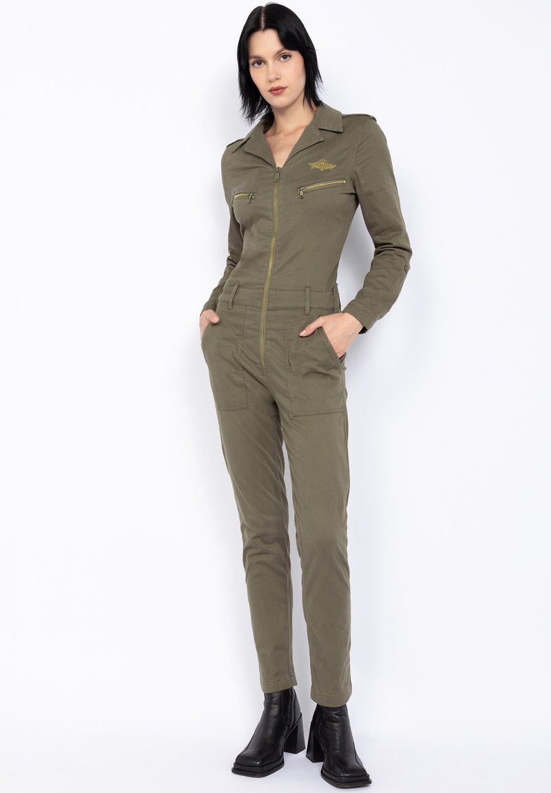 Jumpsuit verde oliva in tessuto di cotone con tasche con zip e colletto. Presenta un design aderente e gambe affusolate, abbinato a stivali neri con tacco.