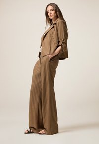 Brunrandig kostym med en kort jacka som har sidofickor och vida byxor. Tyget verkar vara strukturerat. Bärs med svarta sandaler.