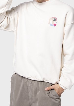 Person trägt einen weißen Pullover mit einem kleinen bunten abstrakten Pflanzendesign auf der Brust, kombiniert mit grauer Hose.
