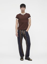 Jeune homme debout, une main dans la poche, portant un t-shirt marron et blanc, un pantalon rayé et des chaussures noires avec des bouts dorés.