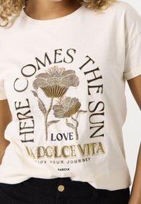 Camiseta de algodón crema con estampados florales metálicos y texto que dice "AQUÍ VIENE EL SOL, AMOR LA DOLCE VITA," con mangas cortas.