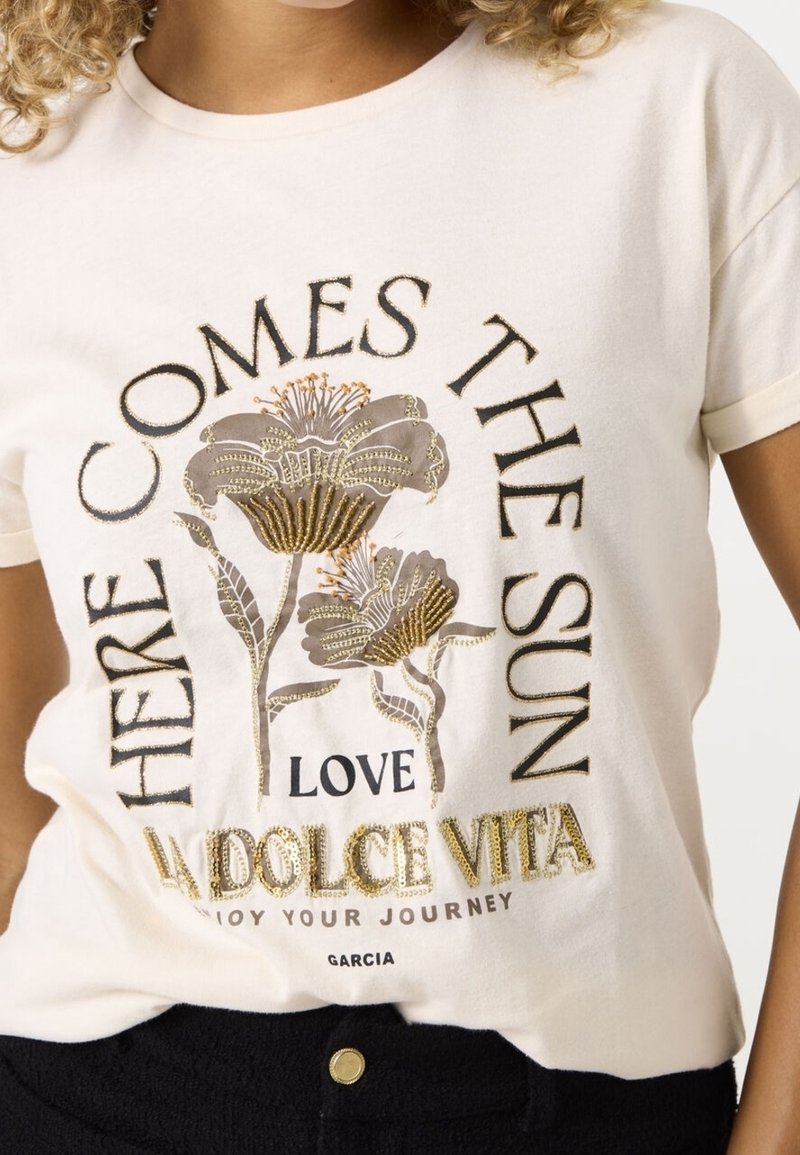 Camiseta de algodón crema con estampados florales metálicos y texto que dice "AQUÍ VIENE EL SOL, AMOR LA DOLCE VITA," con mangas cortas.