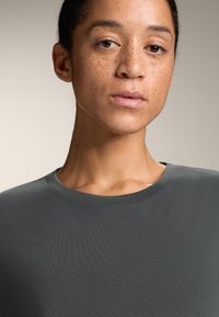 Cămașă gri cu mânecă scurtă și decolteu rotund, fabricată dintr-un material moale și neted. Prezintă o textură discretă cu dungi și cusături îngrijite.