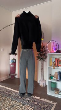 Osoba drží čierny rolák, na sebe má sivé nohavice a čierne topánky, v miestnosti s viditeľným skateboardom, stromom, knižnicou a kobercom.