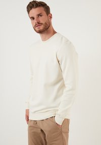 Crème-kleurige sweatshirt met lange mouwen, gemaakt van een textuurstof. Heeft een geribde ronde halslijn en manchetten, gecombineerd met beige broeken.