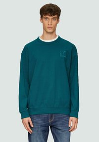 Teal sweatshirt met lange mouwen, ribbelmanchetten en een rechthoekig logo in de linkerbovenhoek, gecombineerd met blauwe jeans. Zachte textuur.