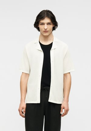 Persona joven con cabello corto y ondulado, vistiendo una camisa blanca de manga corta abierta sobre un top negro y pantalones negros con cordón, de pie frente a un fondo liso.