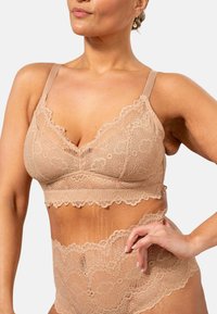 Korsetti pitsistä pehmeässä beigen värissä, jossa on aaltoilevat reunat, varustettuna säädettävillä olkaimilla ja joustavalla vyötärönojalla. Yhdistelmäkorkeat pitsiset alushousut täydentävät settiä.
