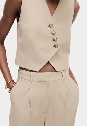 Beżowa bezrękawnikowa krótka kamizelka na cztery guziki z muszli żółwia, zestawiona z beżowymi plisowanymi spodniami z wysokim stanem i złotymi bransoletkami.