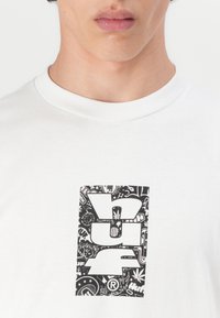 Biele bavlnené tričko s výrazným čiernym grafickým dizajnom s detailnými prvkami, vrátane symbolov a vzorov v logu.