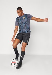 Camiseta de fútbol Puma en gris oscuro con patrón azul, pantalones cortos negros y calcetas negras. El jugador equilibra un balón de fútbol sobre un pie.