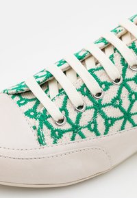 Gros plan sur une chaussure de sport blanche avec une broderie géométrique verte et des lacets blancs passés dans des œillets métalliques.