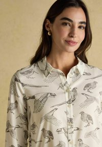 Young woman with long dark hair wears a cream buttoned shirt featuring detailed black bird illustrations and small gold hoop earrings.