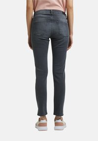 Persona vistiendo jeans ajustados de color gris oscuro y zapatillas beige con acentos rosas, de pie, de espaldas, sobre un fondo claro y liso.