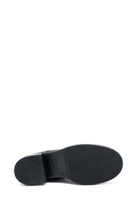 Suela de zapato negra con una superficie de goma texturizada, forma redondeada y tacón plano. Presenta una parte elevada cerca del tacón con detalles de logotipo.