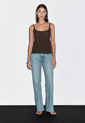 Bruine aansluitende tanktop met dunne bandjes, gecombineerd met lichtblauwe flared jeans. Model staat op een zwarte ondergrond, recht vooruit kijkend.