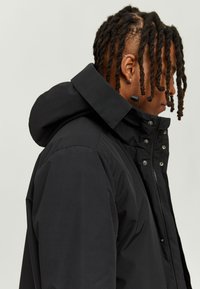 Black nylon jacket with a high collar, adjustable hood, and snap-button details, featuring a smooth texture and sleek design.