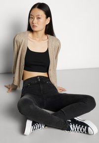 Cardigã de malha bege com mangas bufantes, top crop preto, jeans pretos de cintura alta e sapatilhas pretas com solas brancas, posição sentada.