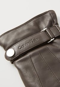 Barna bőrkesztyű sima felülettel, ezüst színű snap gombbal és "Calvin Klein" logóval domborítva ellátott pánttal. Látható varrás részletek.