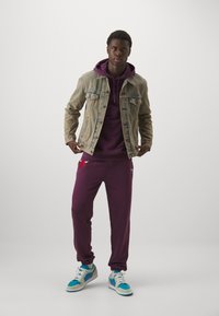 Giacca di jeans sopra un hoodie viola, abbinata a pantaloni della tuta viola con un logo, e sneakers turchesi con accenti grigi.
