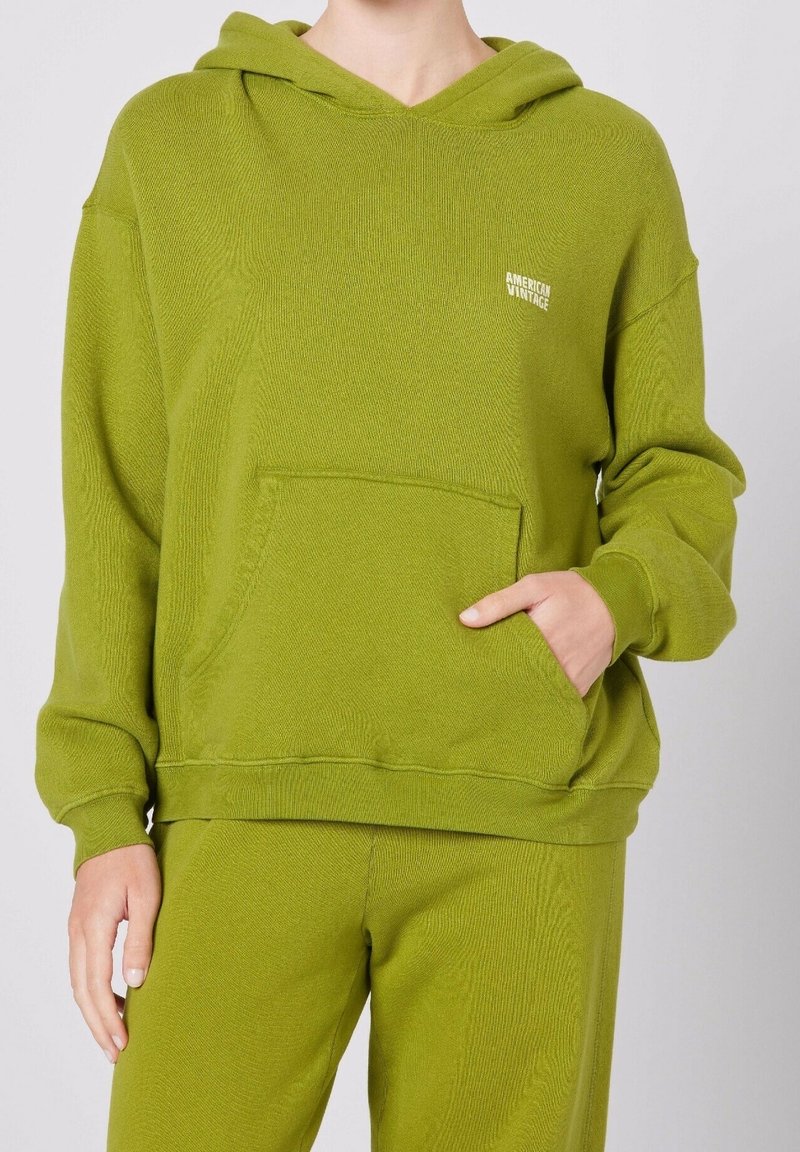 Felpa verde con finitura testurizzata, dotata di tasca frontale e cappuccio regolabile con lacci. Piccolo logo bianco sul petto. Abbinata a pantaloni verdi.