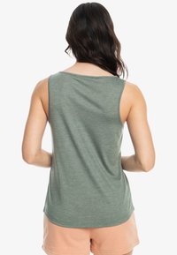 Mujer con pelo largo y oscuro, vestida con una blusa sin mangas verde y pantalones cortos melocotón, de pie de espaldas a la cámara contra un fondo blanco.