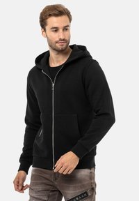 Hoodie noir zippé en mélange de coton, doté d'une capuche doublée, de poches avant et de poignets côtelés. Texture lisse, coupe décontractée.