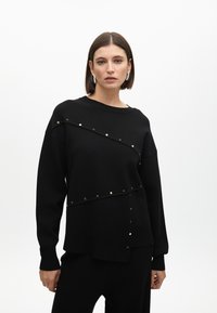 Suéter negro de corte oversized, que presenta tachuelas doradas diagonales en la parte delantera y trasera, mangas largas y una tela texturizada.