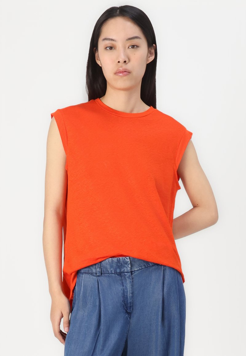 T-shirt sans manches orange en matière douce, avec un col rond, une coupe décontractée et un ourlet arrière légèrement plus long. Associé à un pantalon bleu.