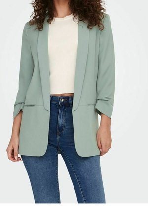 Kvinde iført lys grøn åben blazer over hvid cropped top og blå højttaljede jeans, stående mod en hvid baggrund.