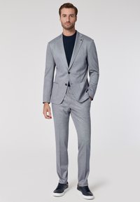 Completo grigio su misura con finitura texturizzata, giacca con un solo bottone e due tasche frontali. Abbinato a una t-shirt blu navy a girocollo e sneakers.