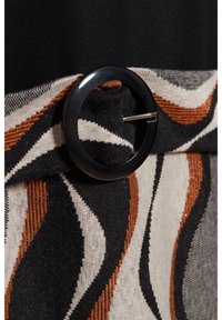 Bälte med en stor svart cirkulär spänne, som har ett mönstrat tyg i svarta, krämfärgade och rostfärgade vågor. Strukturerat och vävt material.