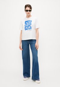 Witte T-shirt met blauwe bloemenillustratie en tekst, gecombineerd met wijde blauwe denim jeans en witte sneakers met paarse accenten.