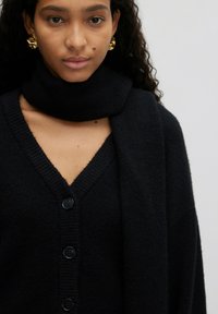 Zwarte gebreide cardigan met een diepe V-hals, voorzien van vijf knopen, gecombineerd met een bijpassende sjaal die om de nek is gedrapeerd. Gelaagd, comfortabel materiaal.
